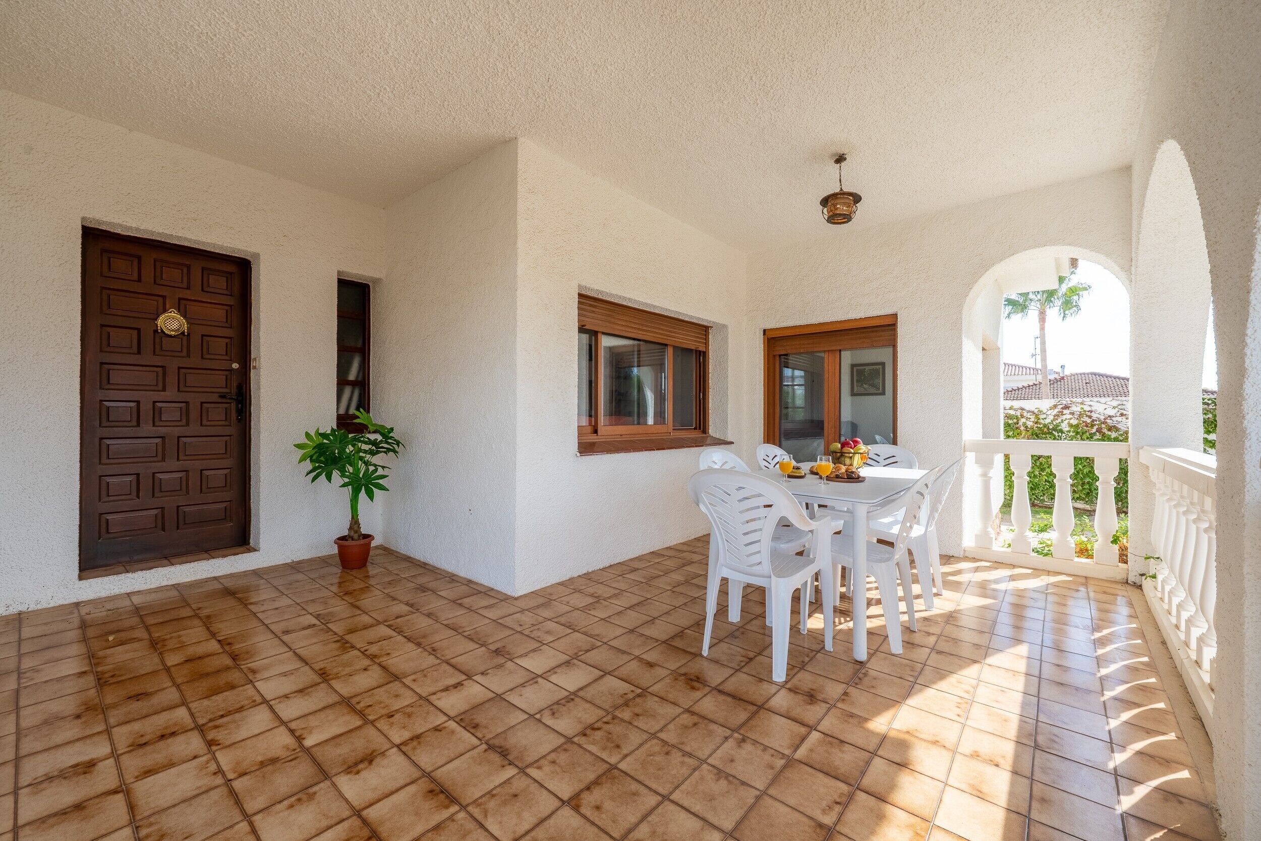 Un patio couvert avec un sol carrelé, une porte en bois, une plante en pot, une table avec des chaises et une rambarde blanche.