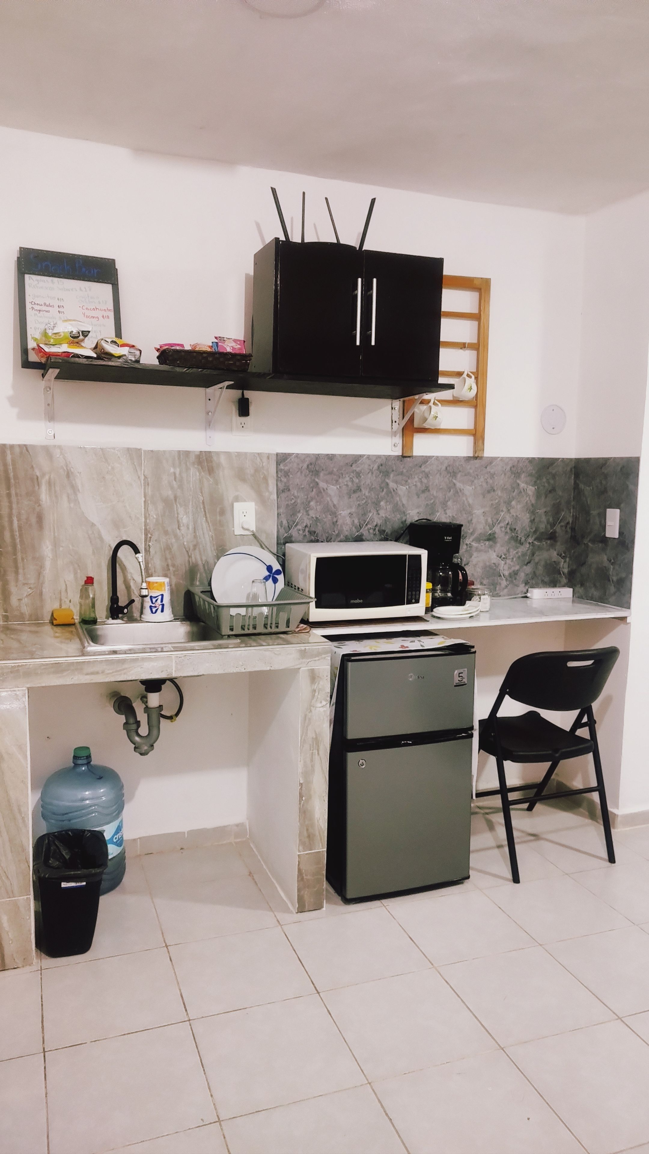 A compact kitchen with a microwave, a small refrigerator, a sink, and a shelf with food items.