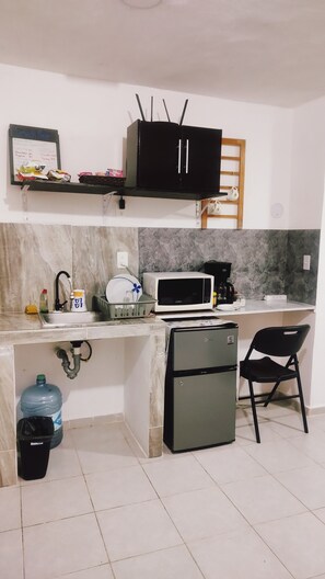 A compact kitchen with a microwave, a small refrigerator, a sink, and a shelf with food items.