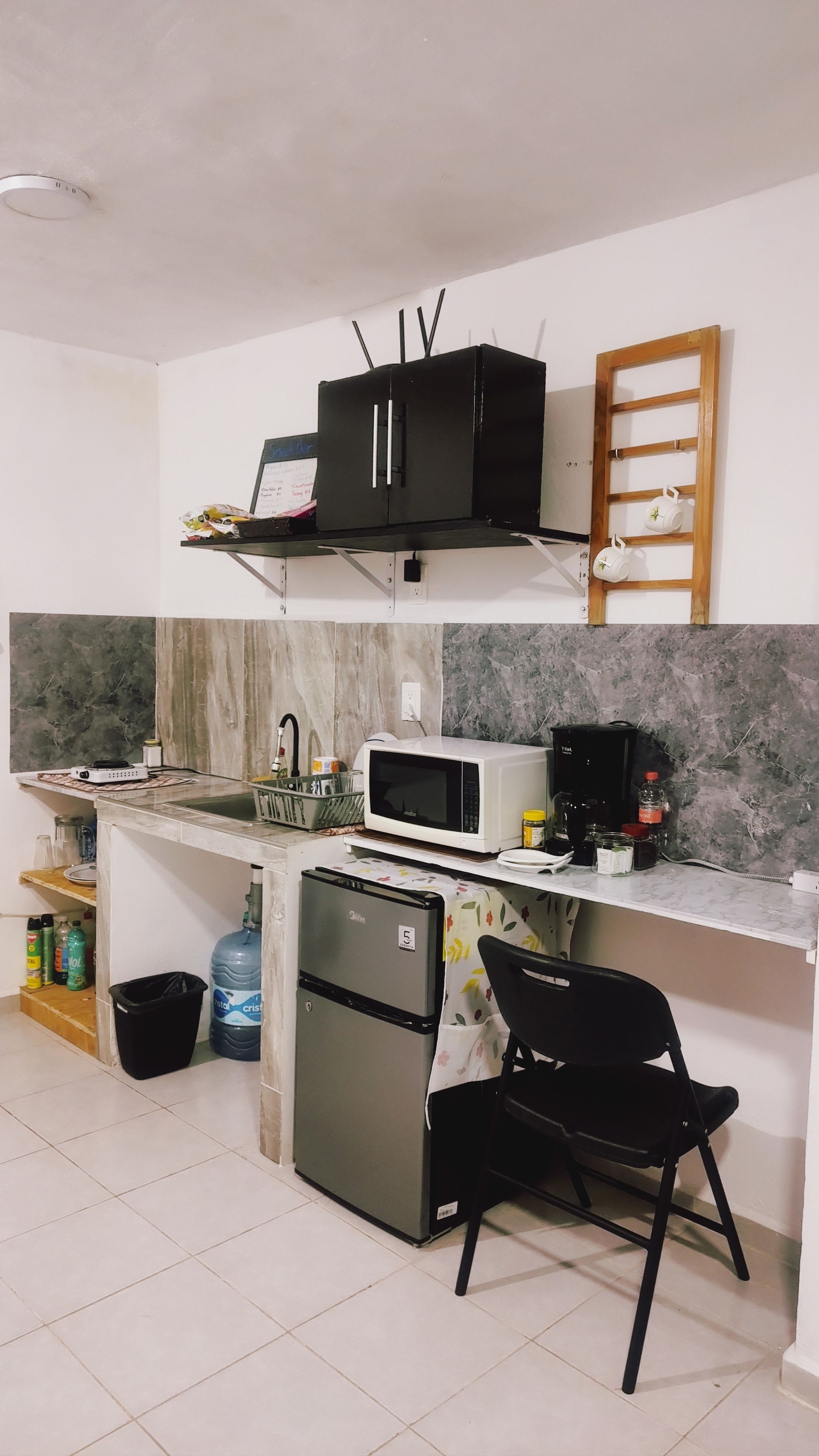 Cocina compacta con microondas, un refrigerador pequeño, un lavamanos y una silla negra.