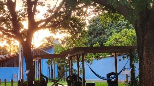 A hammock under a wooden pergola in a garden area with a blue building in the background. - Pousada Bom Jardim (Nobres)