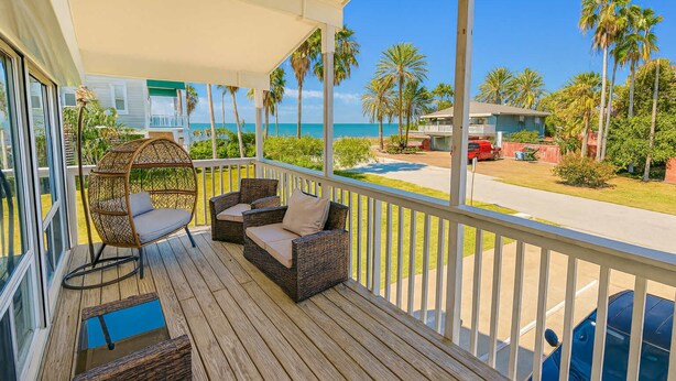 Een houten terras met rieten meubilair, een hangstoel en uitzicht op het strand en palmbomen.
