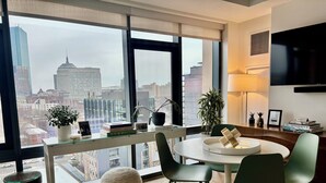 Ett modernt vardagsrum med utsikt över staden, inrett med ett runt bord, gröna stolar, ett soffbord i glas och ett stort fönster med utsikt över stadens silhuett.