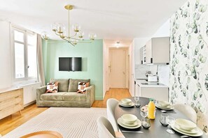 Ein modernes Wohnzimmer mit einer Couch, einem Esstisch für vier Personen, einem an der Wand montierten Fernseher und einem durch eine offene Tür einsehbarer Küchenbereich.