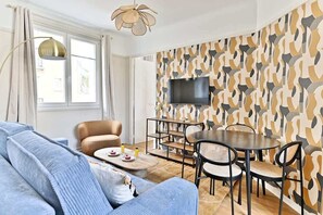 A modern living room with a sofa, a dining table with chairs, a TV mounted on a patterned wall, and a window with curtains. - Calm and light in the heart of Neuilly (Neuilly-sur-Seine)