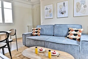 A living room with a blue sofa, two patterned cushions, a coffee table with drinks and plates, and a dining area with chairs and a table. - Calm and light in the heart of Neuilly (Neuilly-sur-Seine)