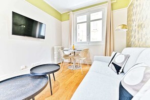 A modern living room with a sofa, a small round table, a flat-screen TV mounted on the wall, and a dining area with a table and chairs. - Charming apartment in Neuilly-sur-Seine (Neuilly-sur-Seine)