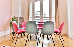 A dining area with a round table and six chairs, a potted plant, and a view of buildings through large windows. - Superb 2-bedroom apartment in Neuilly-sur-Seine (Neuilly-sur-Seine)
