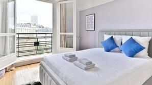 A bedroom with a bed, white bedding, two blue pillows, and a view of buildings through an open window. - Superb 2-bedroom apartment in Neuilly-sur-Seine (Neuilly-sur-Seine)
