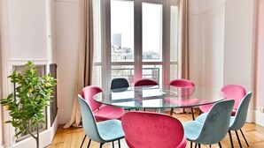 A modern room with a glass table surrounded by colorful chairs, large windows with curtains, and a small plant. - Superb 2-bedroom apartment in Neuilly-sur-Seine (Neuilly-sur-Seine)