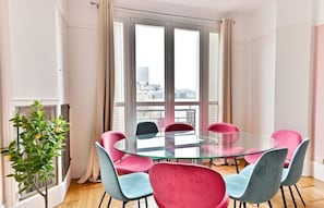 A modern room with a glass table surrounded by colorful chairs, large windows with curtains, and a small plant. - Superb 2-bedroom apartment in Neuilly-sur-Seine (Neuilly-sur-Seine)