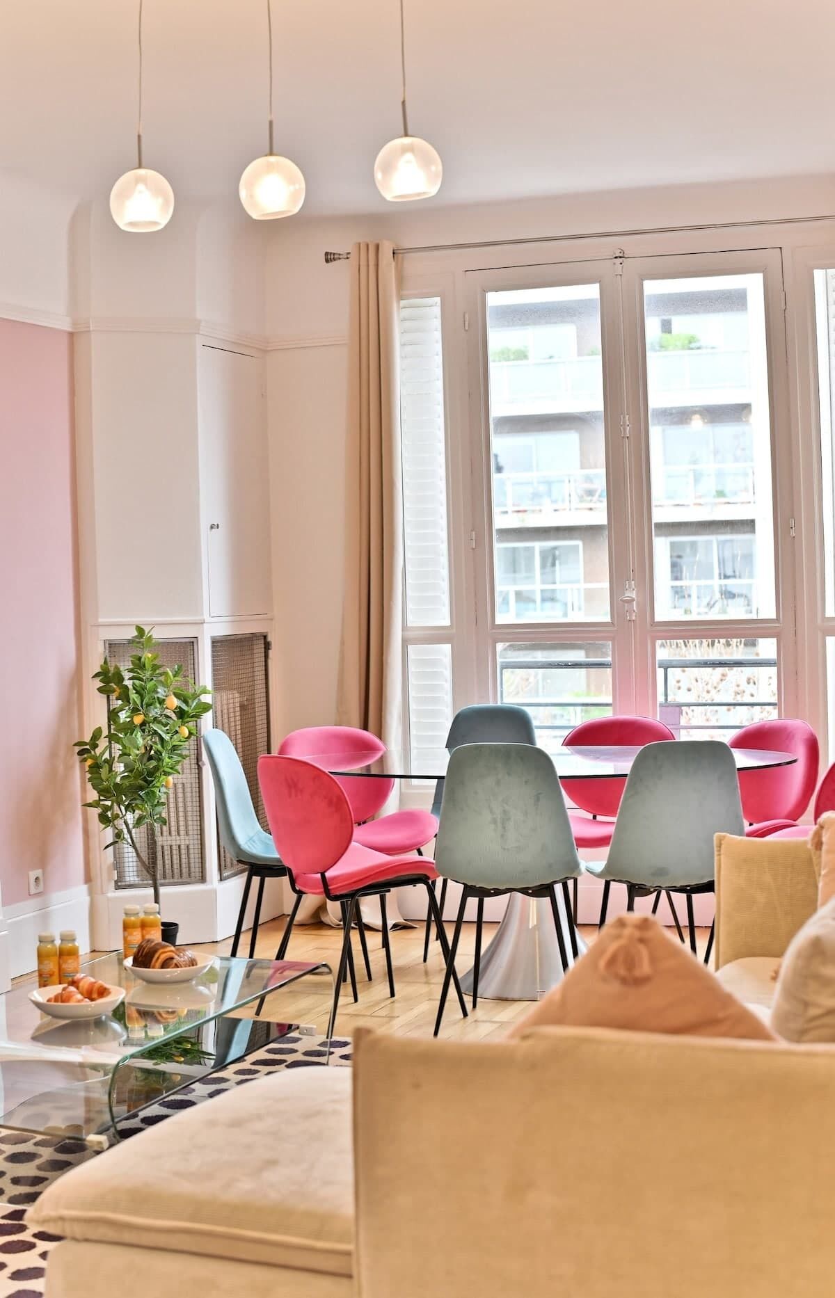 Un salon moderne avec une table basse en verre, un canapé et un coin repas avec des chaises rouges et grises. Une grande fenêtre offre une vue sur les bâtiments à l’extérieur.