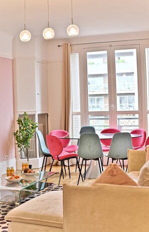 A modern living room with a glass coffee table, a sofa, and a dining area with red and grey chairs. There is a large window with a view of buildings outside. - Superb 2-bedroom apartment in Neuilly-sur-Seine (Neuilly-sur-Seine)