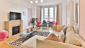 A modern living room with a flat-screen TV, a fireplace, a dining area with pink chairs, and a sofa with patterned cushions. - Superb 2-bedroom apartment in Neuilly-sur-Seine (Neuilly-sur-Seine)