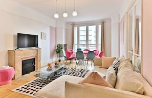 A modern living room with a flat-screen TV, a fireplace, a dining area with pink chairs, and a sofa with patterned cushions. - Superb 2-bedroom apartment in Neuilly-sur-Seine (Neuilly-sur-Seine)