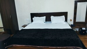 Un lit bien fait, avec du linge de lit blanc et une couvre-lit noire à motifs, une table de chevet en bois avec un téléphone et une petite table à côté, et une armoire en bois sombre.