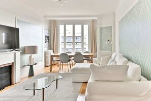 A modern living room with a sofa, a glass coffee table, a TV, and a dining area with chairs and a table. - Beautiful apartment with unobstructed views in Neuilly (Neuilly-sur-Seine)