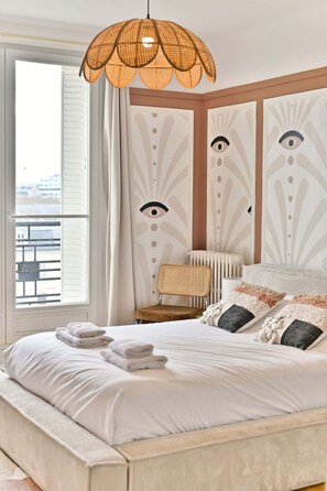 A bedroom with a large bed, a rattan chair, a radiator, and a window with curtains. - Beautiful apartment with unobstructed views in Neuilly (Neuilly-sur-Seine)