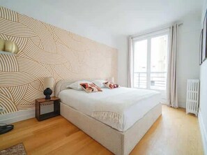 A bedroom with a patterned headboard, a bed with pillows, a bedside table with a lamp, and a window with curtains. - An intimate and bright haven in Neuilly (Neuilly-sur-Seine)