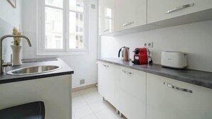 A modern kitchen with white cabinets, a stainless steel sink, a kettle, a toaster, and a window providing natural light. - An intimate and bright haven in Neuilly (Neuilly-sur-Seine)