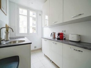 A modern kitchen with white cabinets, a stainless steel sink, a kettle, a toaster, and a window providing natural light. - An intimate and bright haven in Neuilly (Neuilly-sur-Seine)