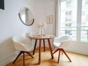 A dining area with a round table, two white chairs, a mirror, and a window with a view of buildings. - An intimate and bright haven in Neuilly (Neuilly-sur-Seine)