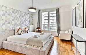 A modern bedroom with a large bed, a sofa, a small table, and a view of the city through the window. - Neuilly-sur-Seine, family cocoon (Neuilly-sur-Seine)