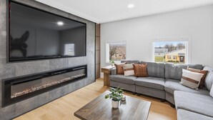 A modern living room with a flat-screen TV, a fireplace, a grey sofa, and a wooden coffee table. - Amazing 5BR Retreat Near Downtown Indy (Indianapolis)