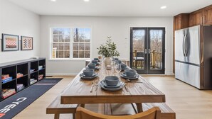 A modern kitchen with a wooden table set for dining, a refrigerator, and a bookshelf with board games. - Amazing 5BR Retreat Near Downtown Indy (Indianapolis)