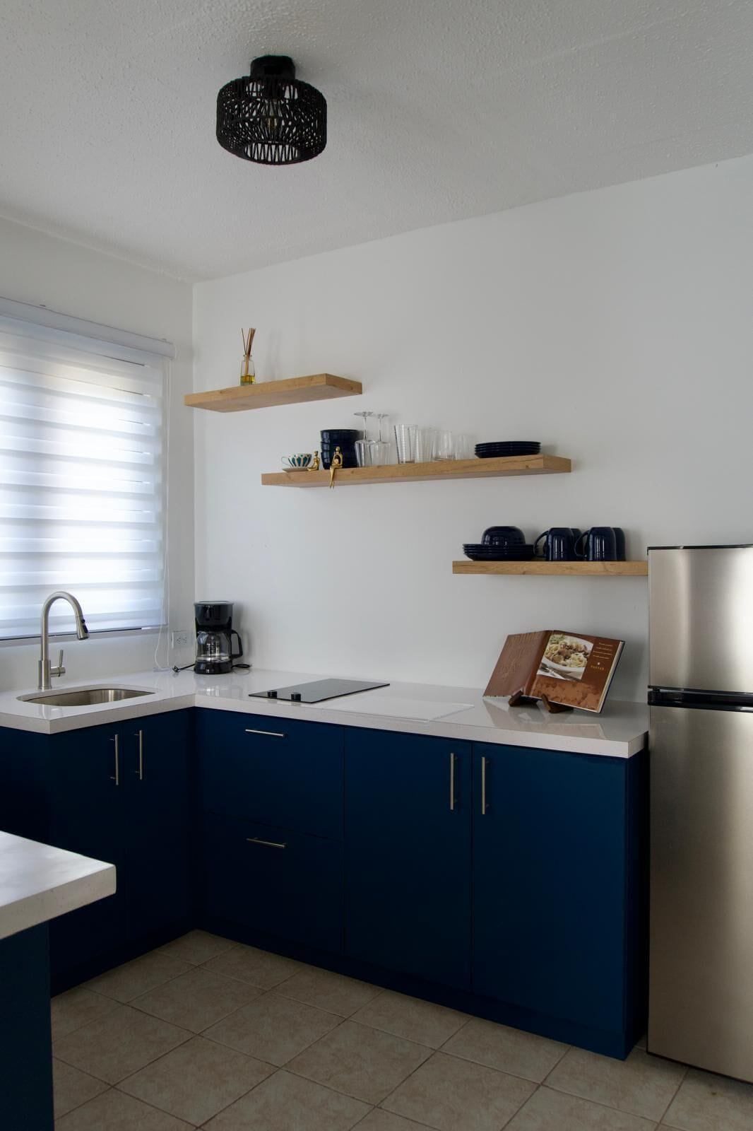 A modern kitchen with dark blue cabinets, white countertops, and stainless steel appliances. There are wooden shelves above the counter holding dishes and a coffee maker.