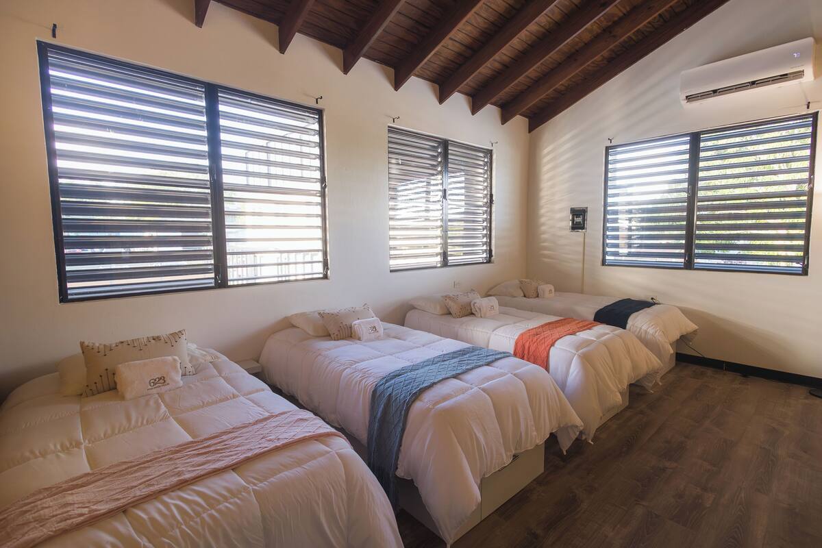 A room with three beds, wooden beams on the ceiling, and large windows with blinds.