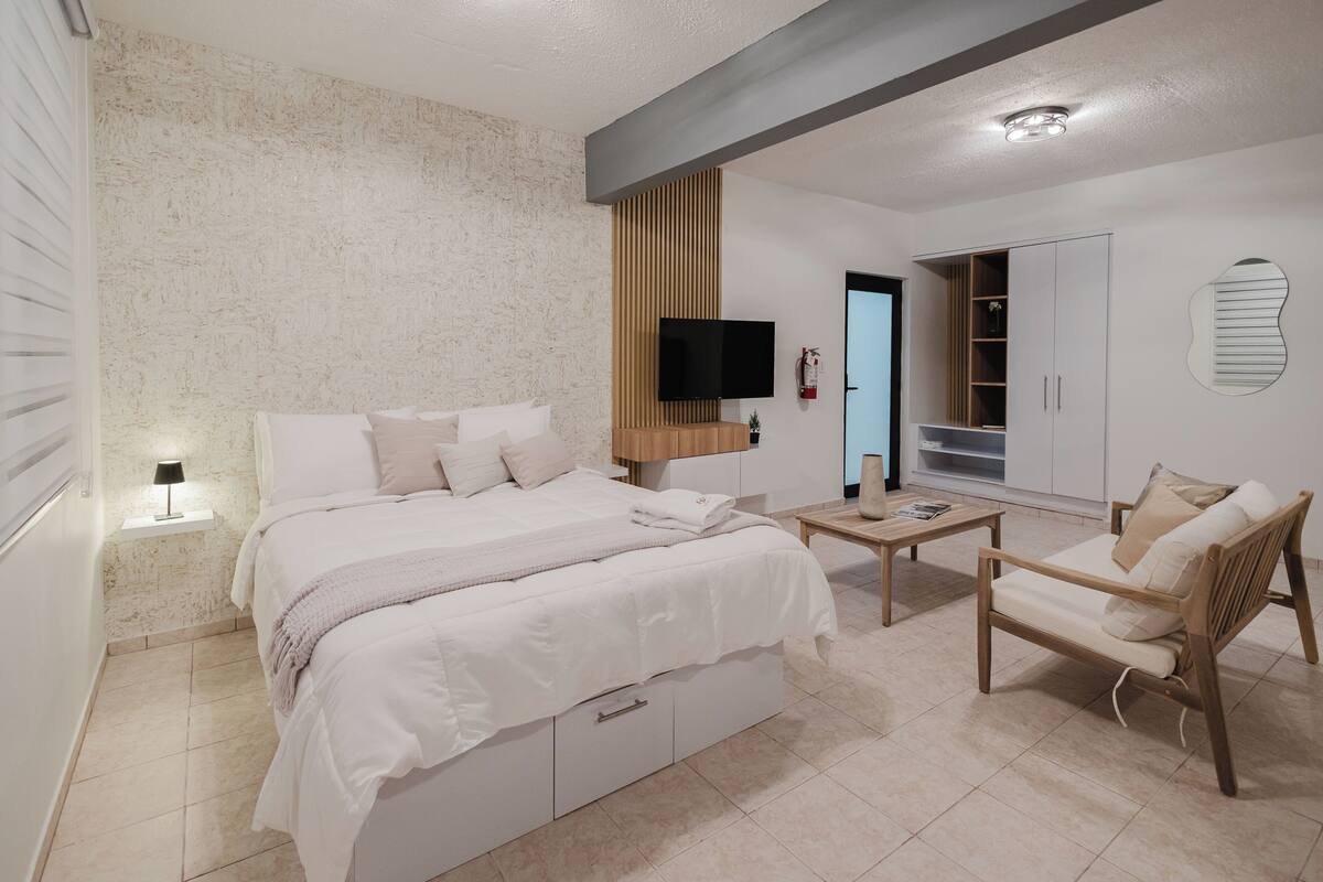 A bedroom with a bed, bedside table, TV, wardrobe, and a seating area with a chair and table.