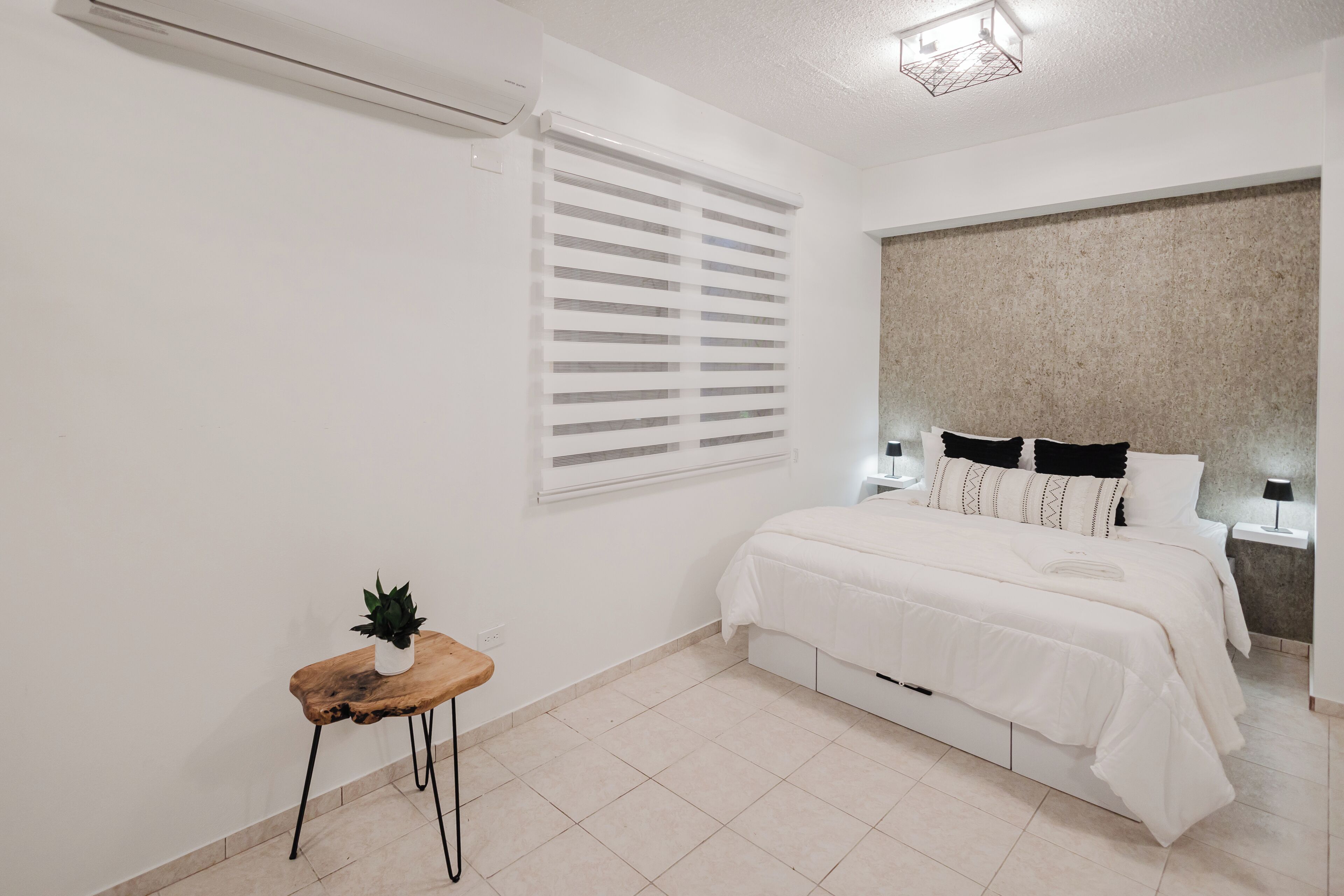 A bedroom with a bed, bedside tables, a potted plant, and a window with blinds.