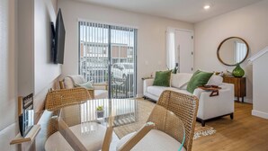 Ett modernt vardagsrum med ett matbord i glas, flätade stolar, en vit soffa med gröna kuddar och ett stort fönster med utsikt över gatan.