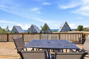 Una terraza de madera con una mesa y sillas de metal, con vistas a casas de playa con techos inclinados y un cielo azul despejado.