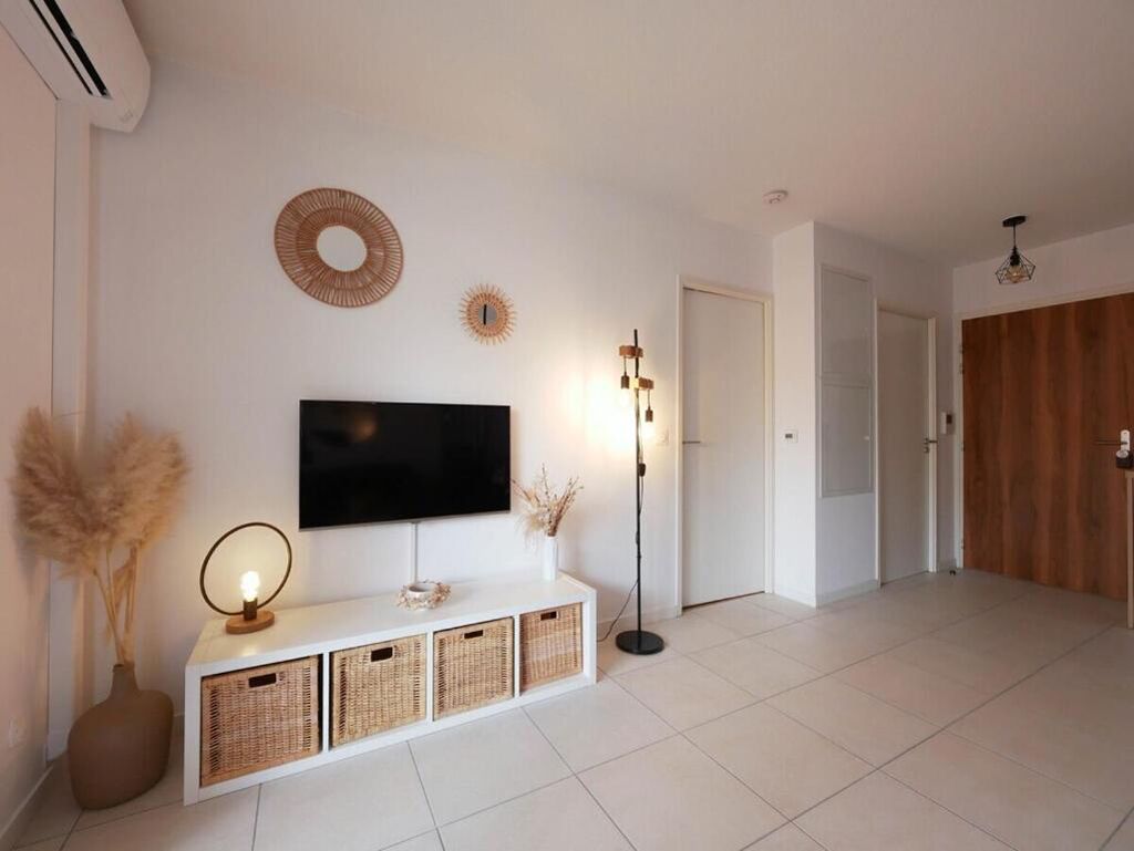Un salon moderne avec un téléviseur  à écran plat fixé au mur, une console blanche agrémentée de paniers en osier, une lampe sur pied et, au fond, une porte en bois.