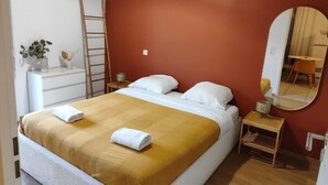 Ein ordentlich bezogenes Bett mit weißer und senffarbener Bettwäsche, ein kleiner Nachttisch mit einer Lampe, ein Spiegel an der Wand und eine Holzleiter, die an der Wand lehnt.