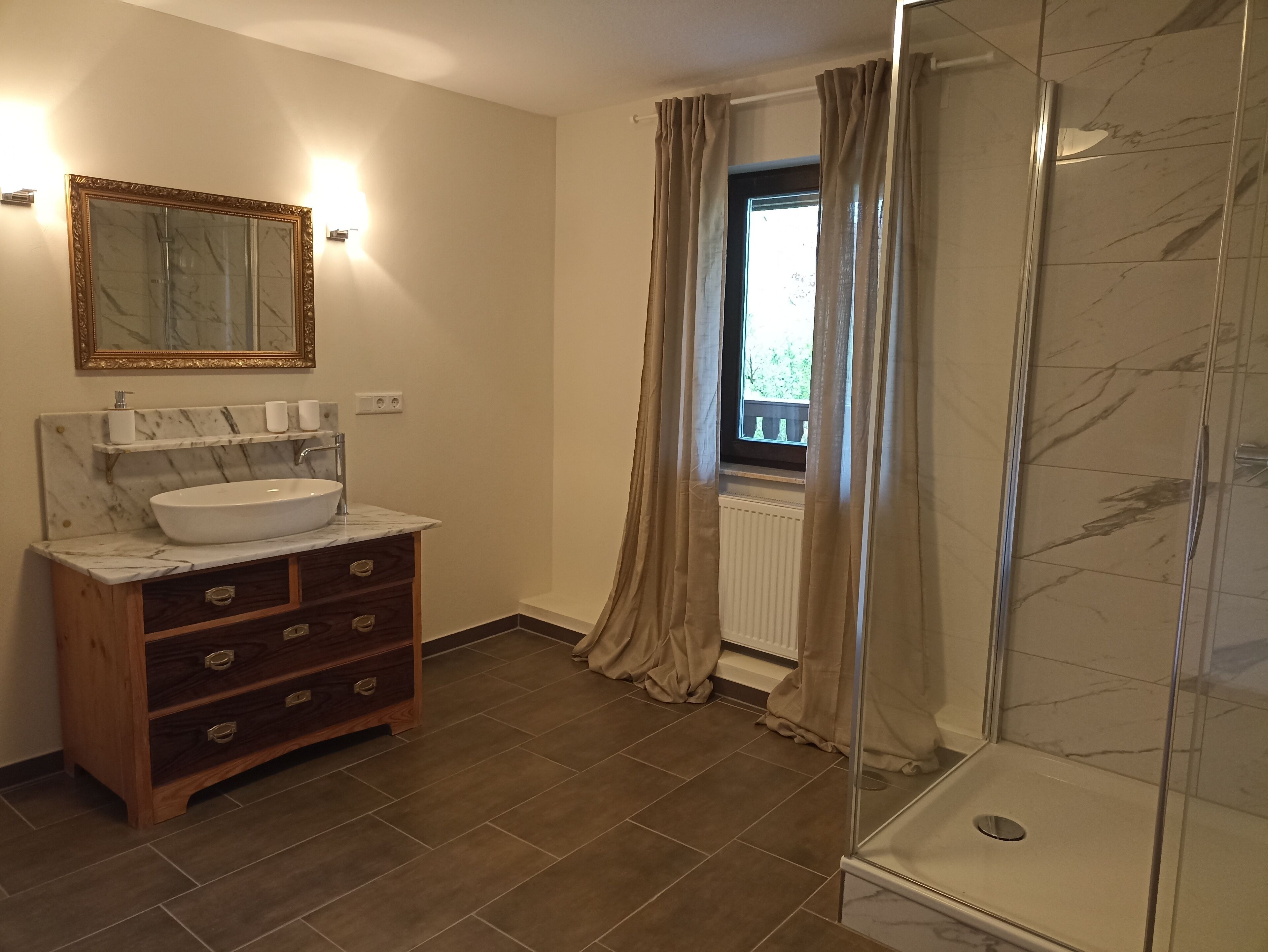 A bathroom with a marble sink, wooden vanity, a glass shower enclosure, and a window with curtains.
