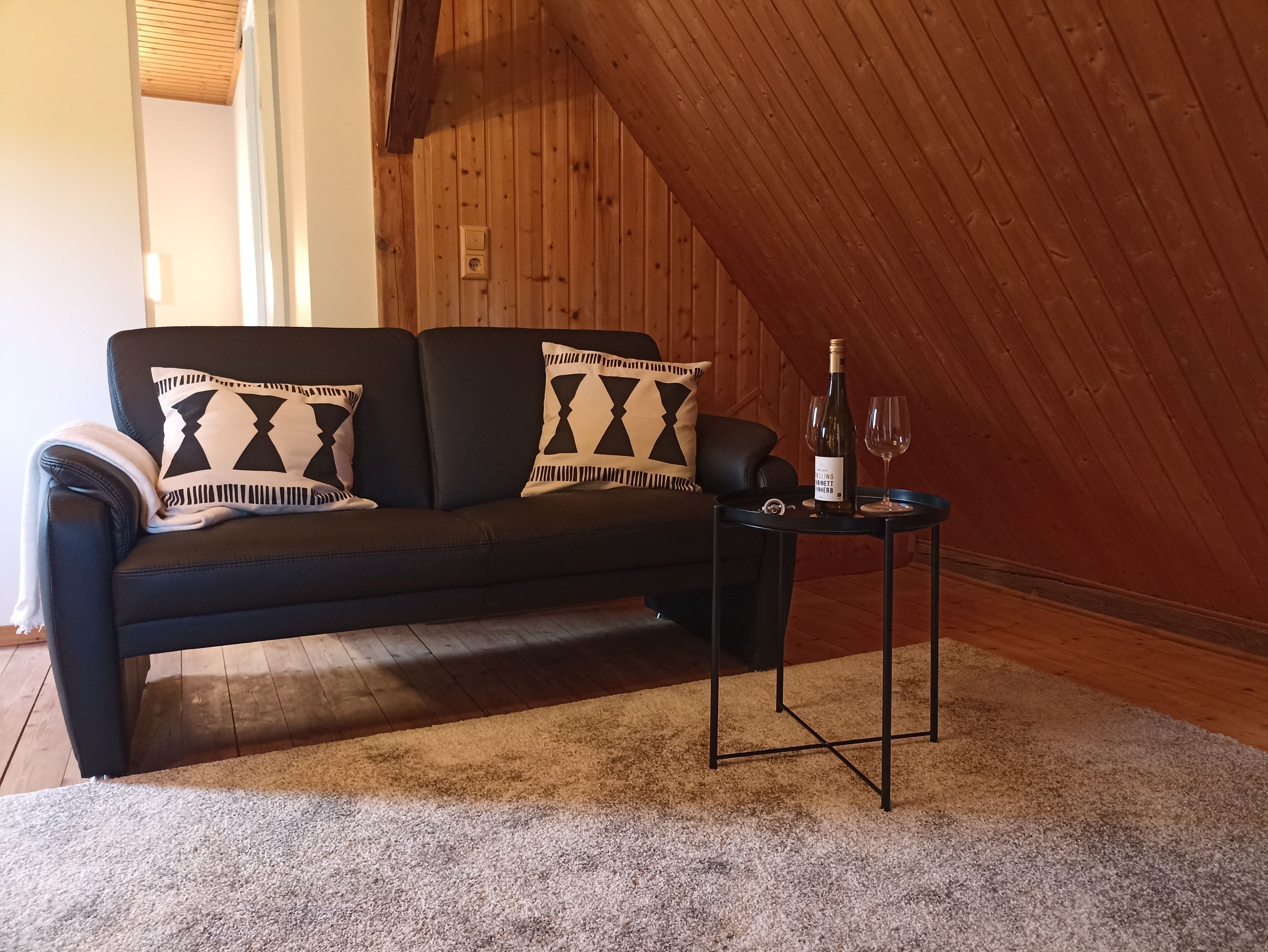 A cozy living room with a dark sofa, patterned cushions, a small table with a bottle and glasses, and a wooden ceiling.