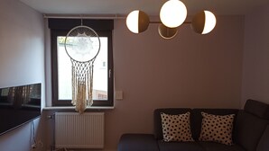 En moderne stue med en mørk sofa, to puder med mønster, en drømmefanger og en usædvanlig loftslampe.