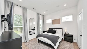 A modern bedroom with a large bed, bedside tables, a mirror, and a television mounted on the wall. - Modern 2BR Townhome Near NRG • Med Center • Downtown w/ Garage (Houston)