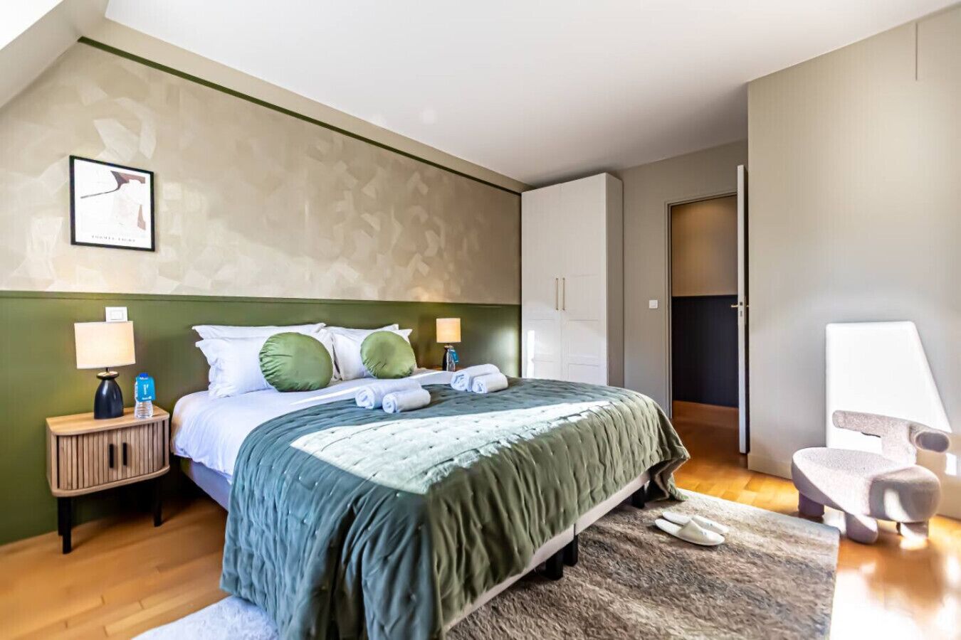 A modern hotel room with a large bed, green and white bedding, a wooden nightstand with a lamp, a small table with a lamp, and a grey rug.