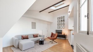 Una sala de estar moderna con un sofá seccional gris, un sillón de cuero marrón y una pequeña mesa redonda. Un tapete con estampado geométrico cubre el suelo de madera. Se observa un televisor montado en la pared y un espejo decorativo hexagonal.