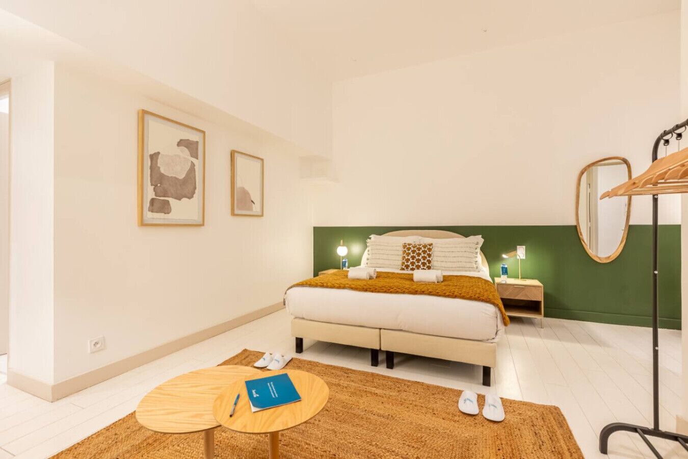Quarto com cama, mesas de cabeceira, espelho, mesa de madeira redonda e um par de chinelos.