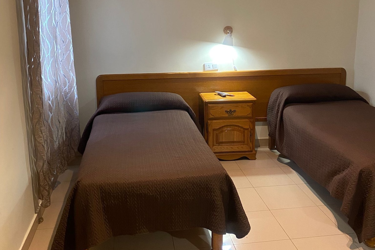 A hotel room with two beds, a nightstand, and a lamp.
