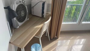 A small, clean room with a wooden desk, two air conditioning units, a white chair, a blue trash bin, and a view of a balcony with greenery. - Hostal Retiro (Madrid)