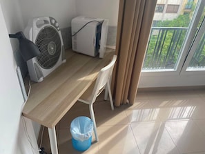 A small, clean room with a wooden desk, two air conditioning units, a white chair, a blue trash bin, and a view of a balcony with greenery. - Hostal Retiro (Madrid)