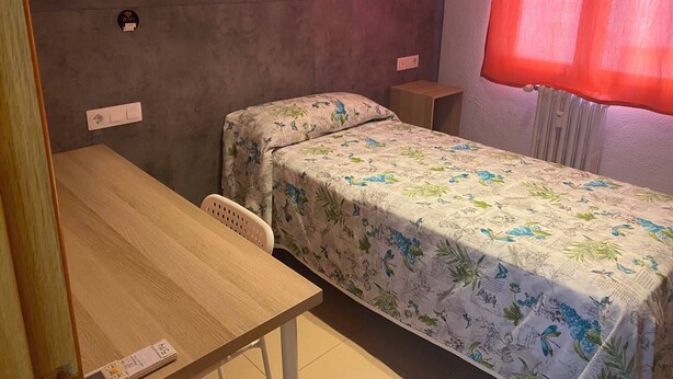 A single bed with a floral bedspread, a wooden desk, a chair, and a window with a red curtain. - Hostal Retiro (Madrid)