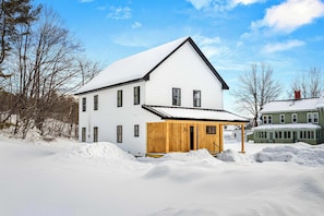 A two-story white house with a wooden porch, surrounded by snow. - Modern Ludlow Home | Walk to Village | 5 Min Okemo (Ludlow)