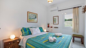 A bedroom with a bed, bedside tables, a hanging light fixture, a window with curtains, and a wall-mounted air conditioning unit. - Entire Cottage in Kerames, Crete – 250 m from the Beach (Agiou Vasileiou)